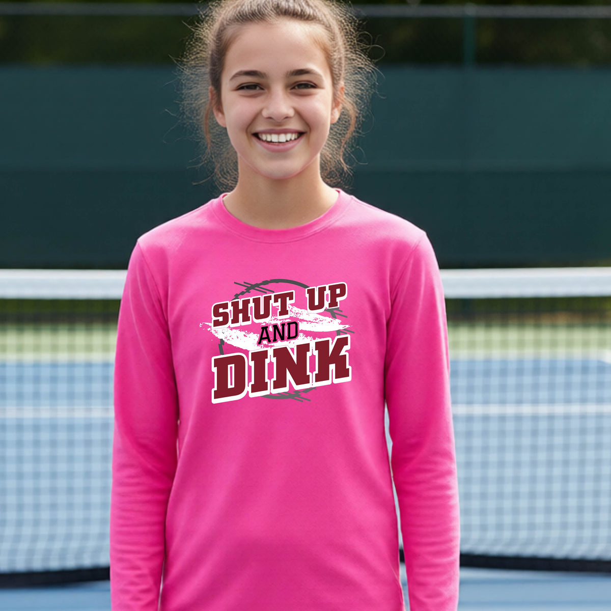 A young person on a pickleball court wearing a pink long-sleeve t-shirt with a white, gray and black graphic of the text:  ‘Shut Up and Dink, standing on a pickleball court.