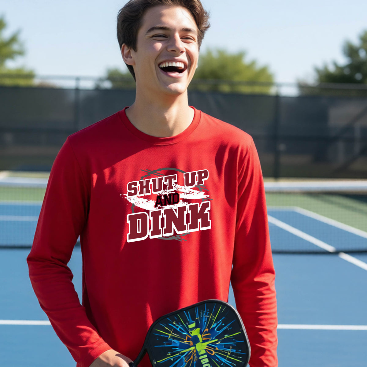 A young person on a pickleball court wearing a red long-sleeve t-shirt with a white, gray and black graphic of the text:  ‘Shut Up and Dink, standing on a pickleball court.
