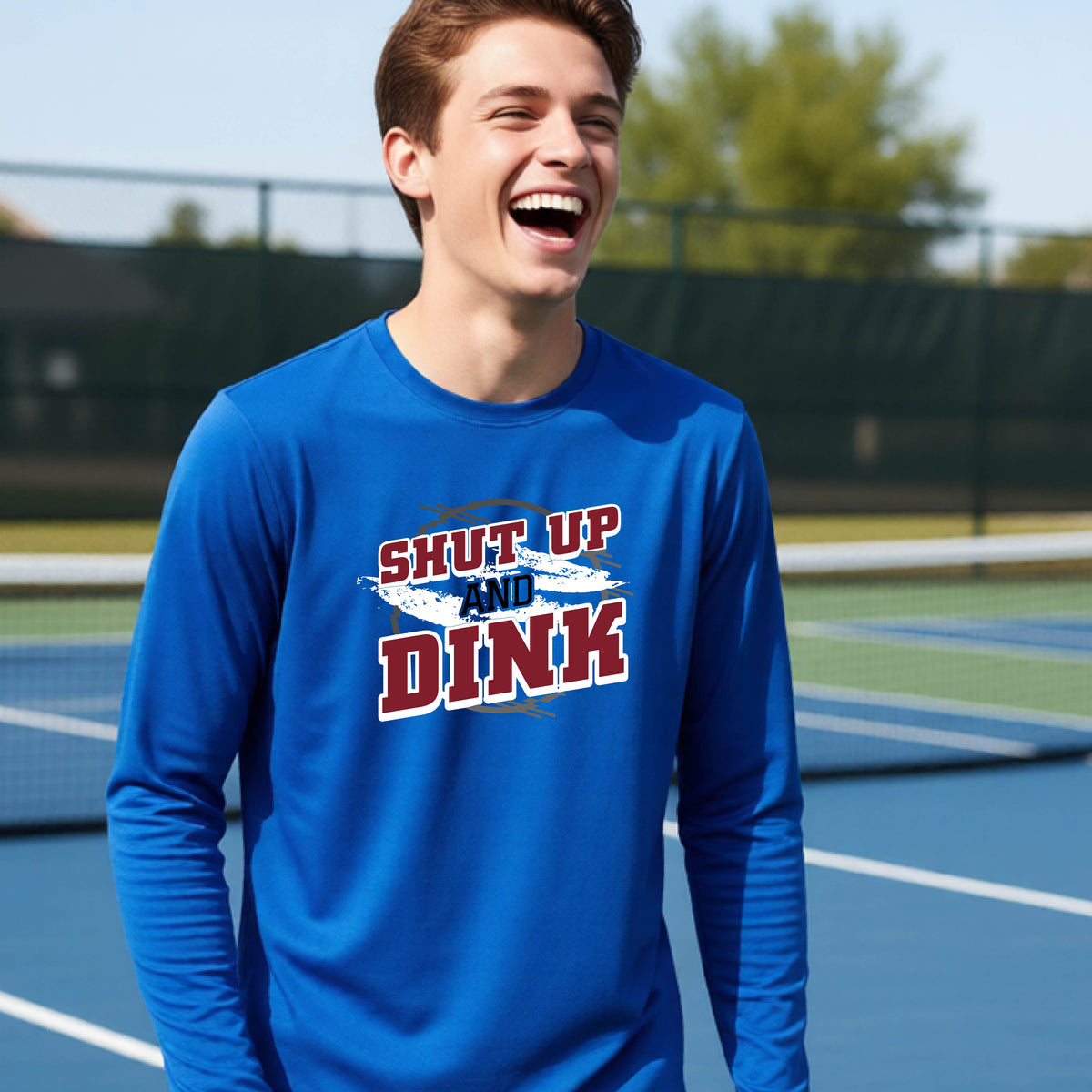 A young person on a pickleball court wearing a royal blue long-sleeve t-shirt with a white, gray and black graphic of the text:  ‘Shut Up and Dink, standing on a pickleball court.