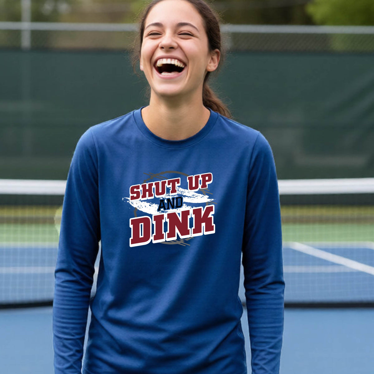 A young person on a pickleball court wearing a royal blue long-sleeve t-shirt with a white, gray and black graphic of the text:  ‘Shut Up and Dink, standing on a pickleball court.