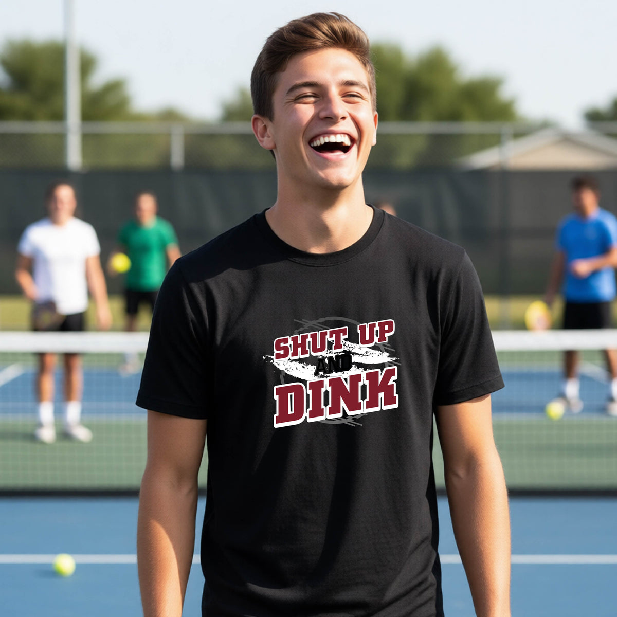 A young person on a pickleball court wearing a black short-sleeve t-shirt with a white, gray and black graphic of the text: ‘Shut Up and Dink, standing on a pickleball court.