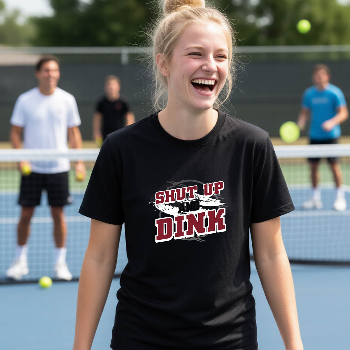 A young person on a pickleball court wearing a black short-sleeve t-shirt with a white, gray and black graphic of the text: ‘Shut Up and Dink, standing on a pickleball court.