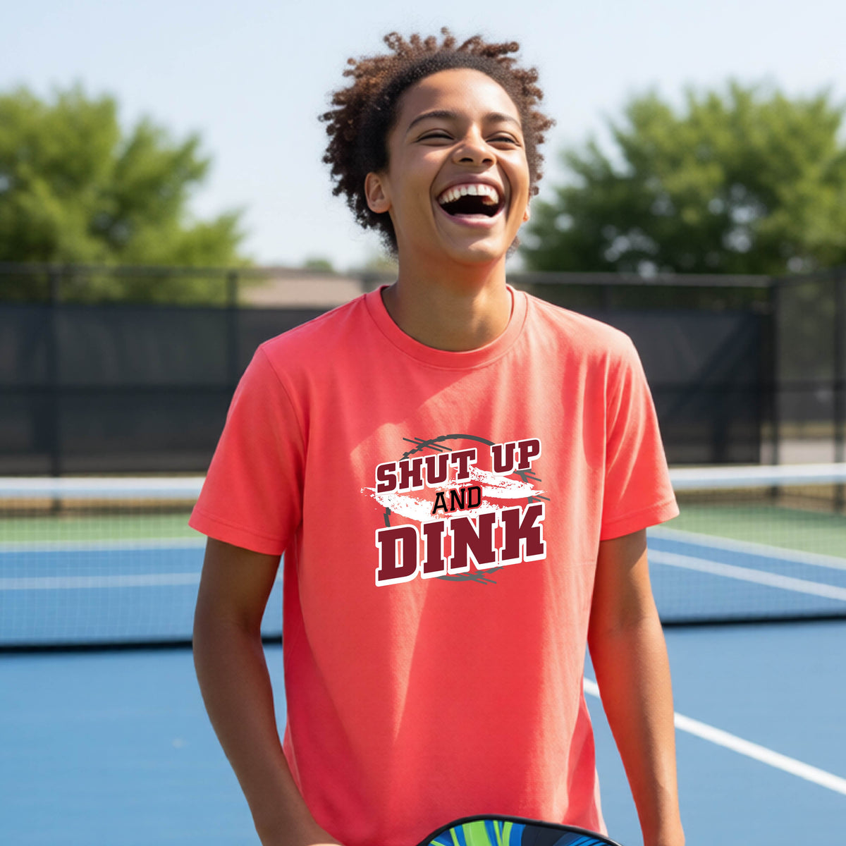 A young person on a pickleball court wearing a bright coral pink short-sleeve t-shirt with a white, gray and black graphic of the text: ‘Shut Up and Dink, standing on a pickleball court.