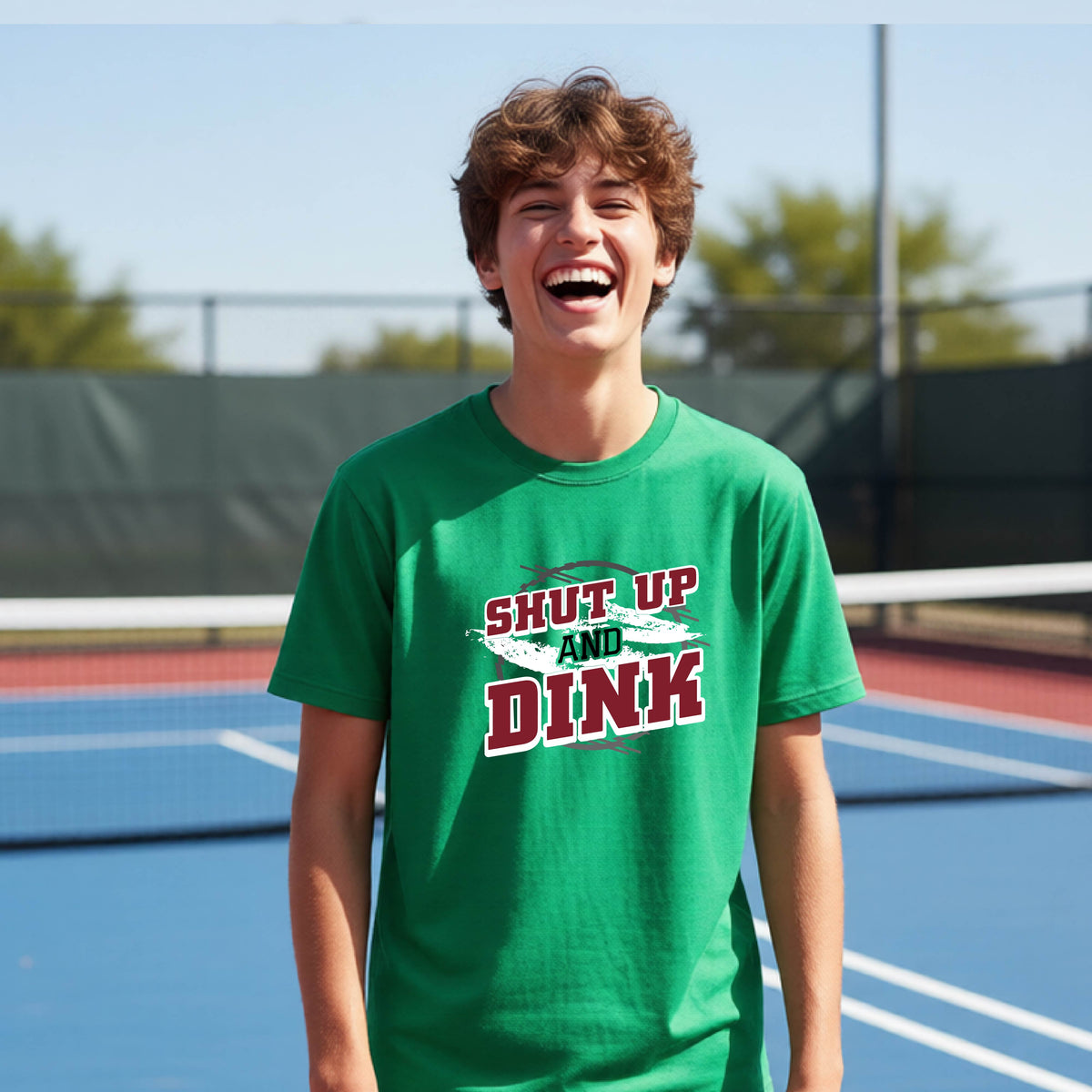 A young person on a pickleball court wearing a green short-sleeve t-shirt with a white, gray and black graphic of the text: ‘Shut Up and Dink, standing on a pickleball court.