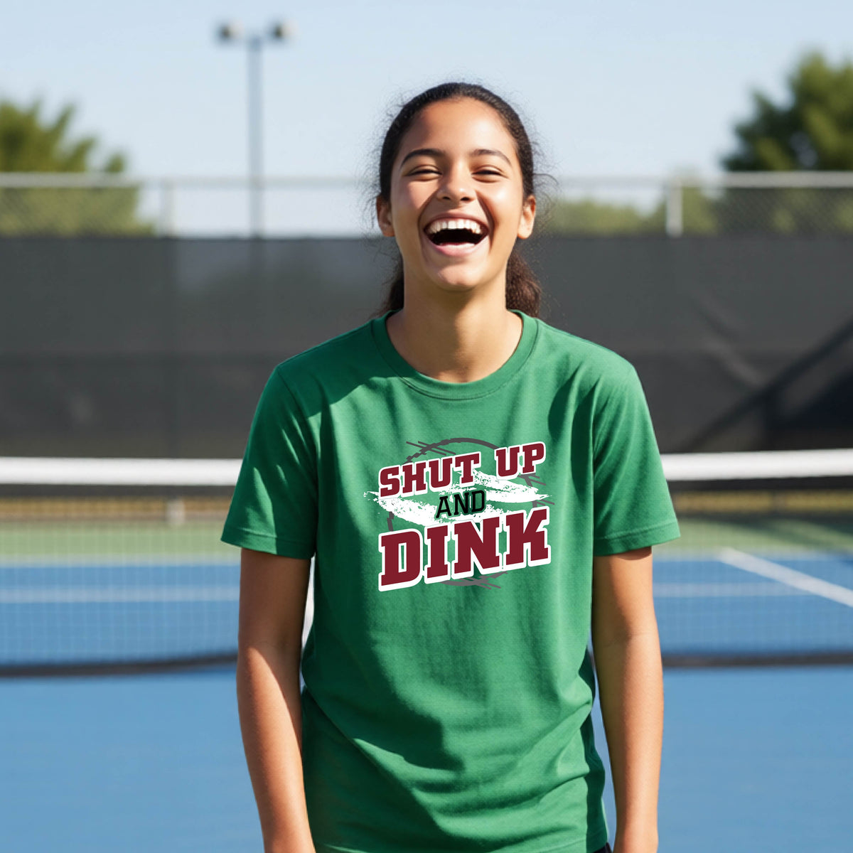 A young person on a pickleball court wearing a green short-sleeve t-shirt with a white, gray and black graphic of the text: ‘Shut Up and Dink, standing on a pickleball court.