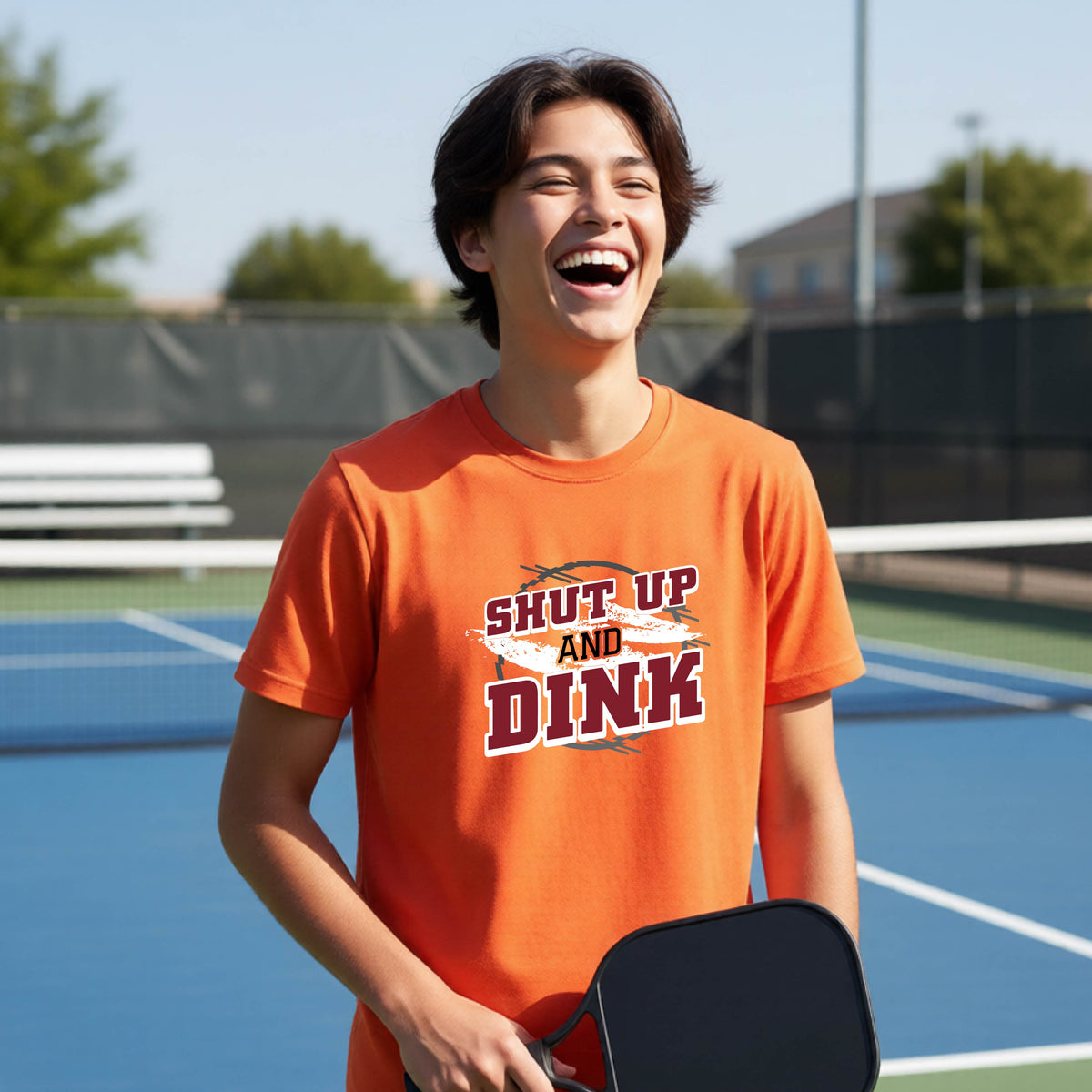 A young person on a pickleball court wearing an orange short-sleeve t-shirt with a white, gray and black graphic of the text: ‘Shut Up and Dink, standing on a pickleball court.