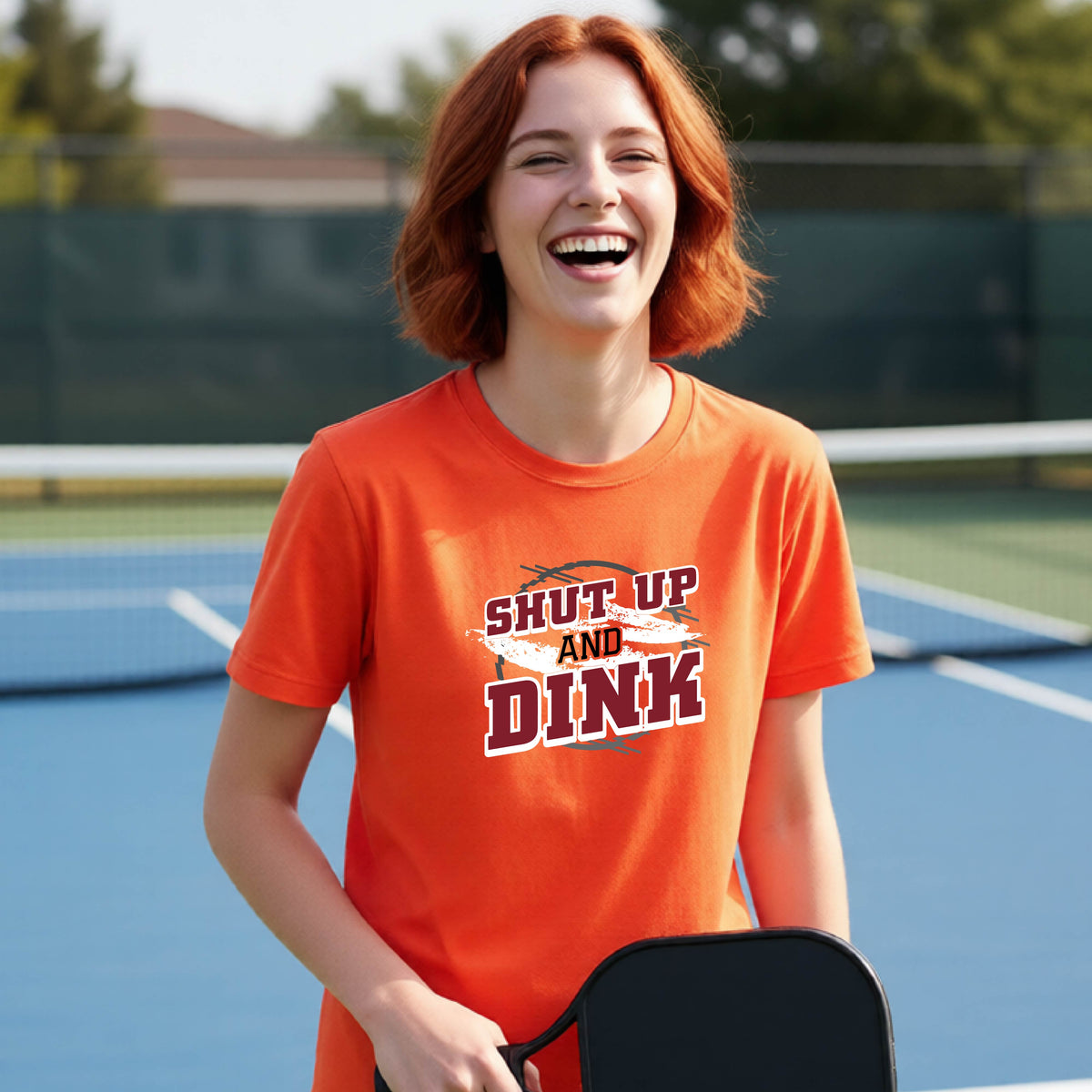 A young person on a pickleball court wearing an orange short-sleeve t-shirt with a white, gray and black graphic of the text: ‘Shut Up and Dink, standing on a pickleball court.