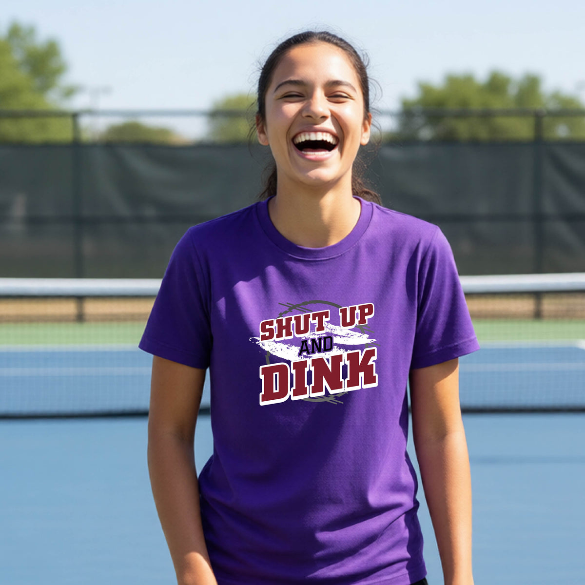 A young person on a pickleball court wearing a purple short-sleeve t-shirt with a white, gray and black graphic of the text: ‘Shut Up and Dink, standing on a pickleball court.