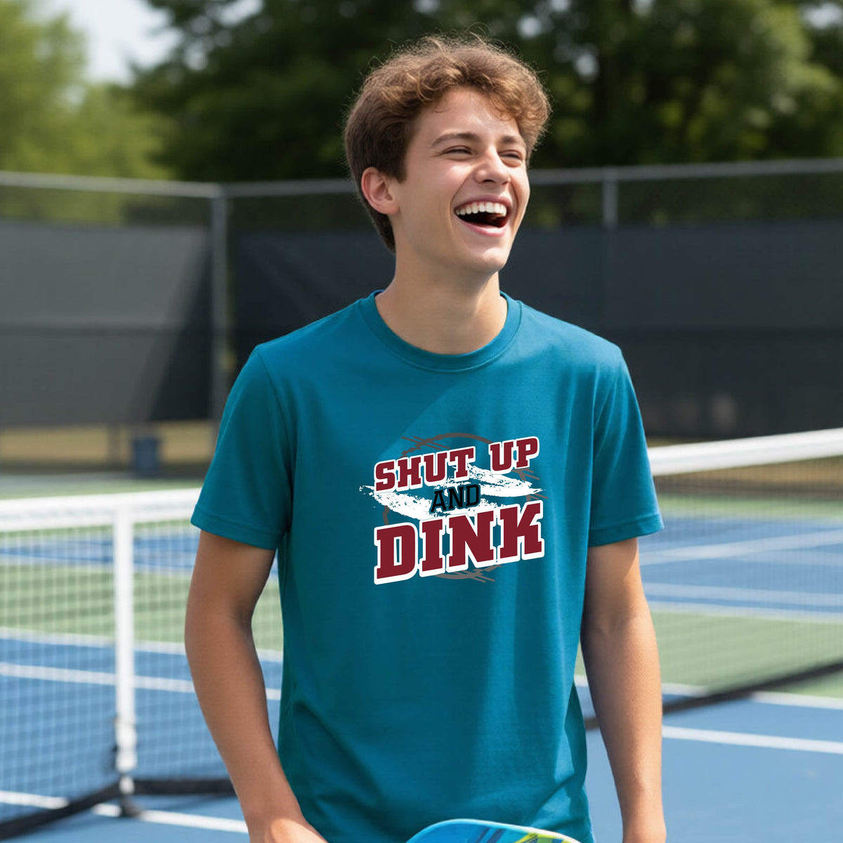 A young person on a pickleball court wearing a teal short-sleeve t-shirt with a white, gray and black graphic of the text: ‘Shut Up and Dink, standing on a pickleball court.