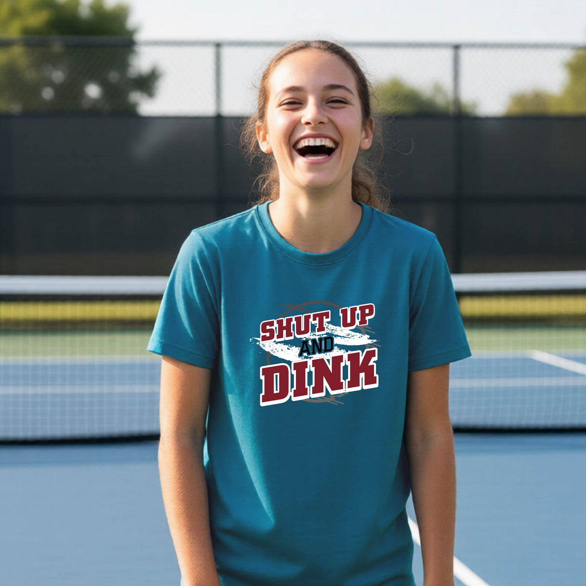 A young person on a pickleball court wearing a teal short-sleeve t-shirt with a white, gray and black graphic of the text: ‘Shut Up and Dink, standing on a pickleball court.