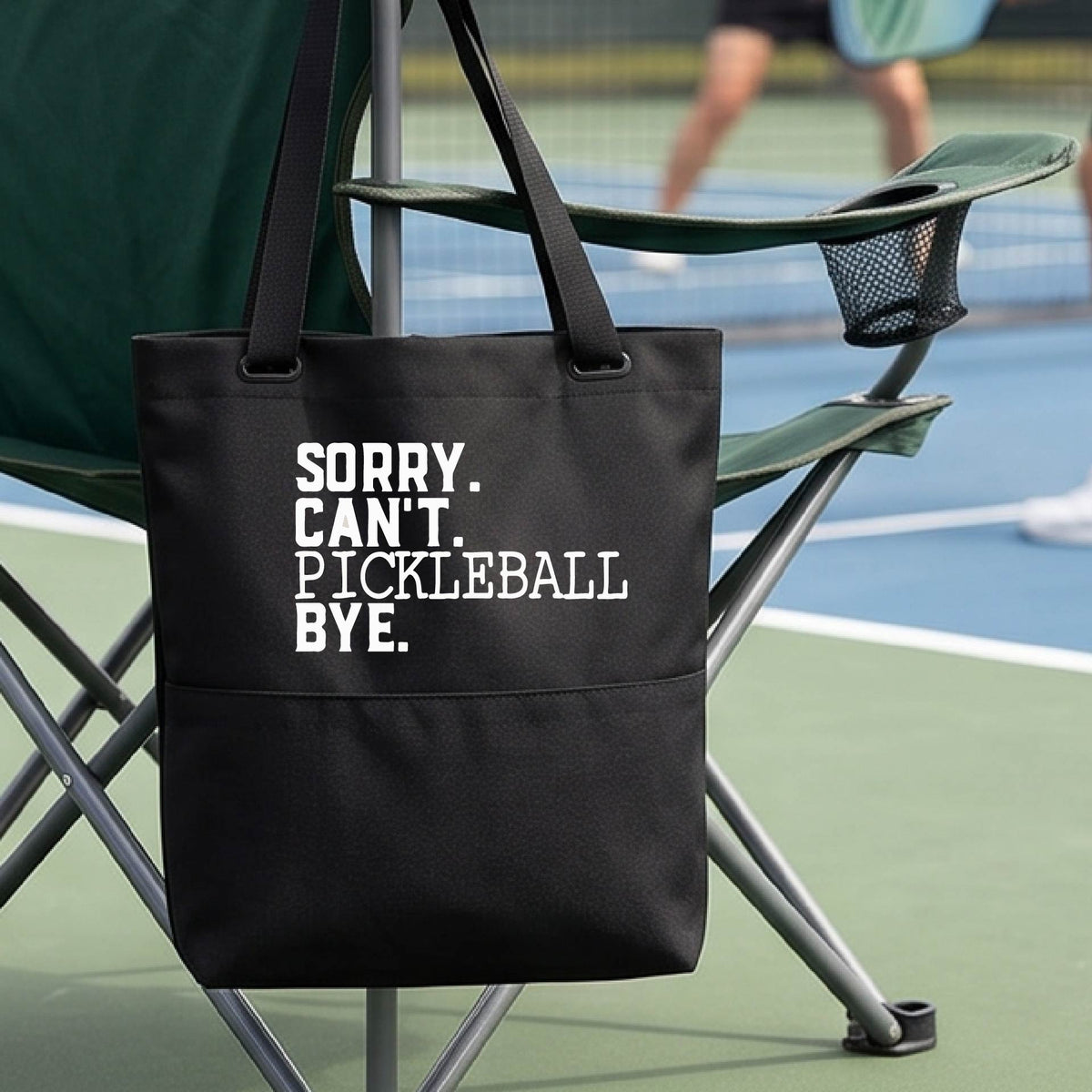 A close up of a black medium size tote bag on a pickleball court with a white graphic of the words  “SORRY. CAN’T. PICKLEBALL BYE” each on a separate line, on the front. 