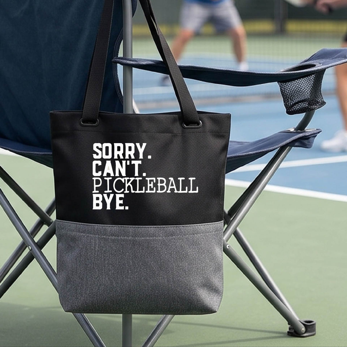 A close up of a grey and black medium size tote bag on a pickleball court with a white graphic of the words  “SORRY. CAN’T. PICKLEBALL BYE” each on a separate line, on the front. 
