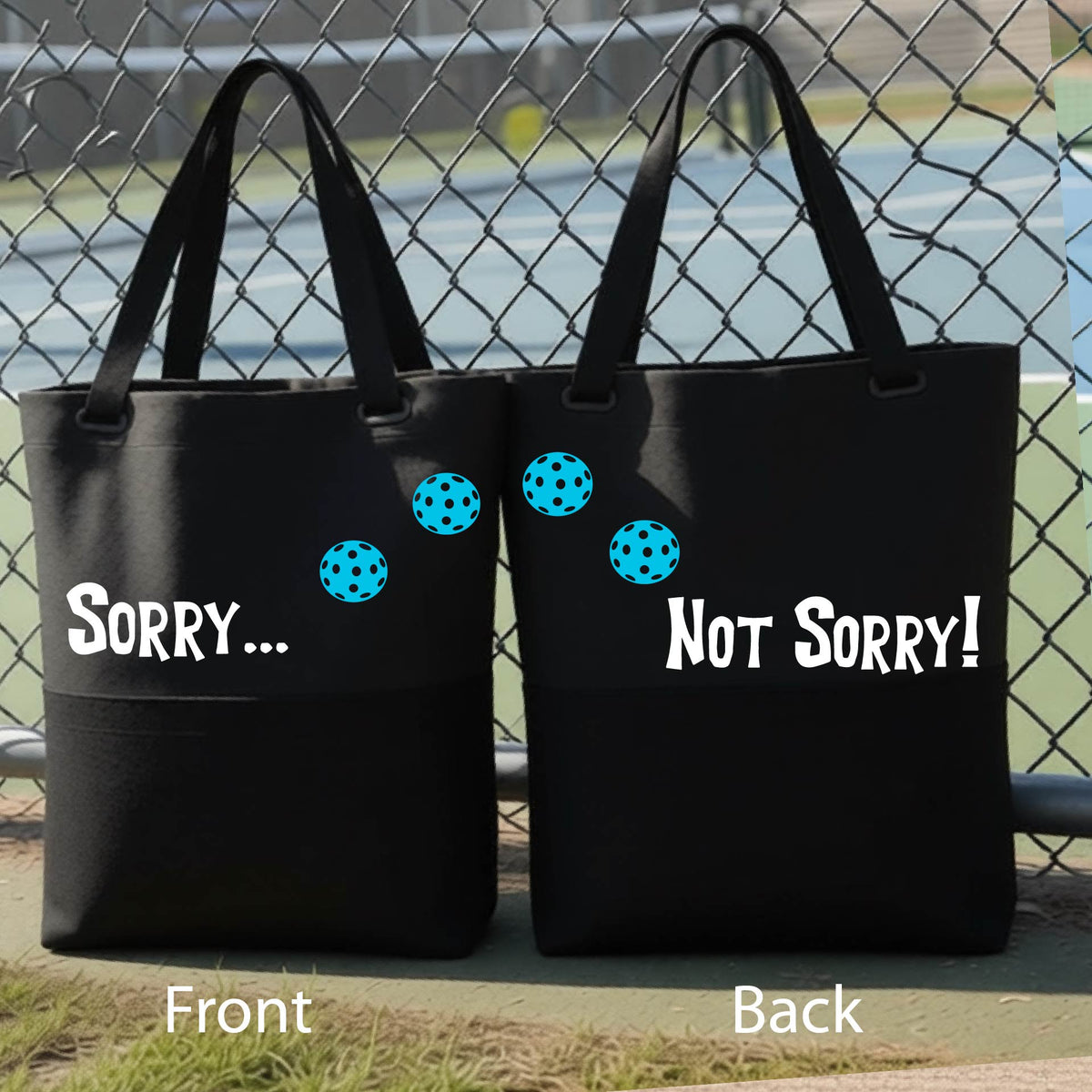 A close-up of a black medium size duffel bag hanging on a fence at a pickleball court, with pickleball motif design on the front and brim and features the playful word “SORRY…” in bold white lettering, accented a colorful pickleball, with the words 'Not sorry" curved underneath on the front of the duffel bag. 