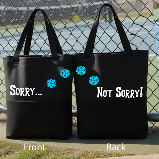 A close-up of a black medium size duffel bag hanging on a fence at a pickleball court, with pickleball motif design on the front and brim and features the playful word “SORRY…” in bold white lettering, accented a colorful pickleball, with the words 'Not sorry" curved underneath on the front of the duffel bag. 