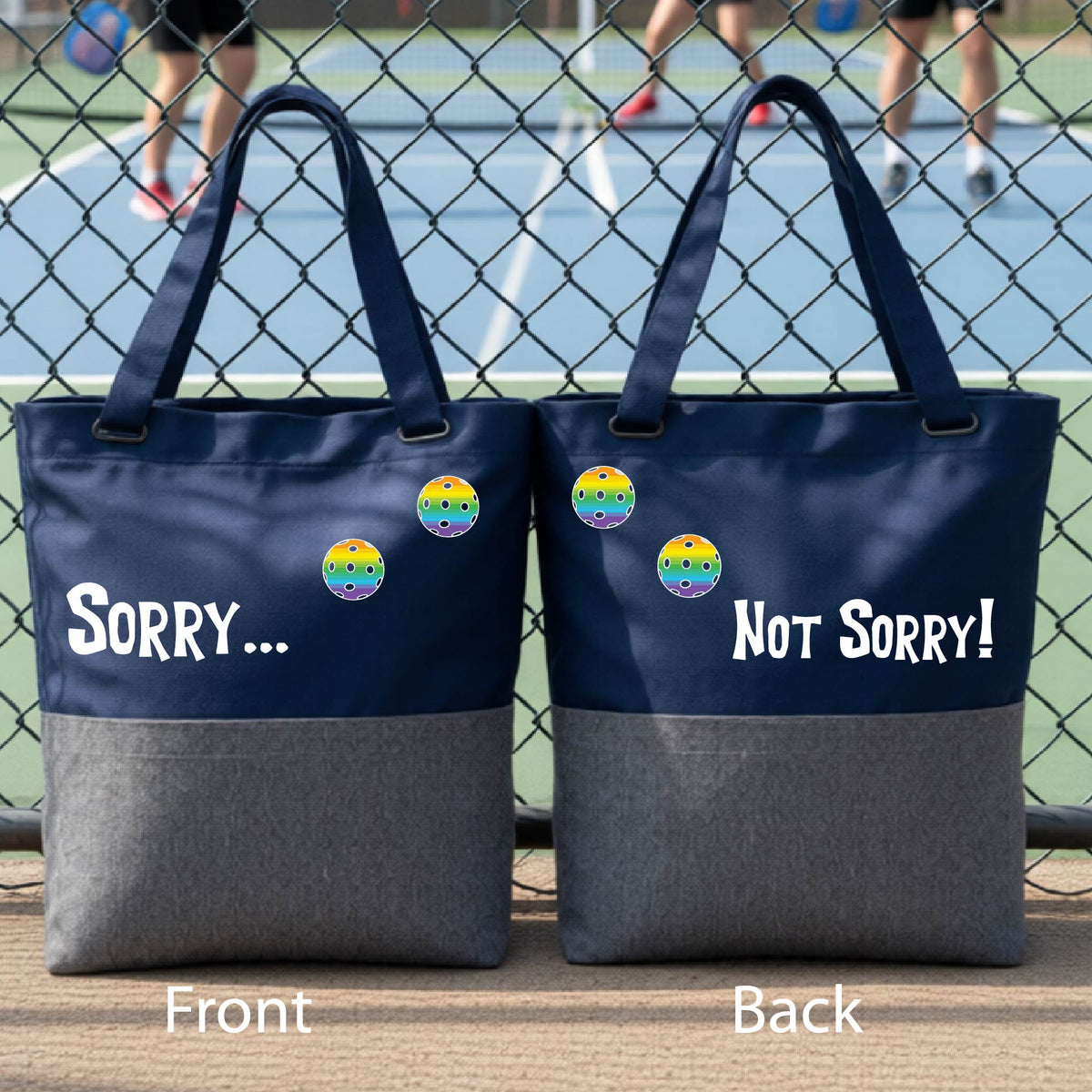 A close-up of a navy blue and gray medium size duffel bag hanging on a fence at a pickleball court, with pickleball motif design on the front and brim and features the playful word “SORRY…” in bold white lettering, accented a colorful pickleball, with the words 'Not sorry" curved underneath on the front of the duffel bag. 