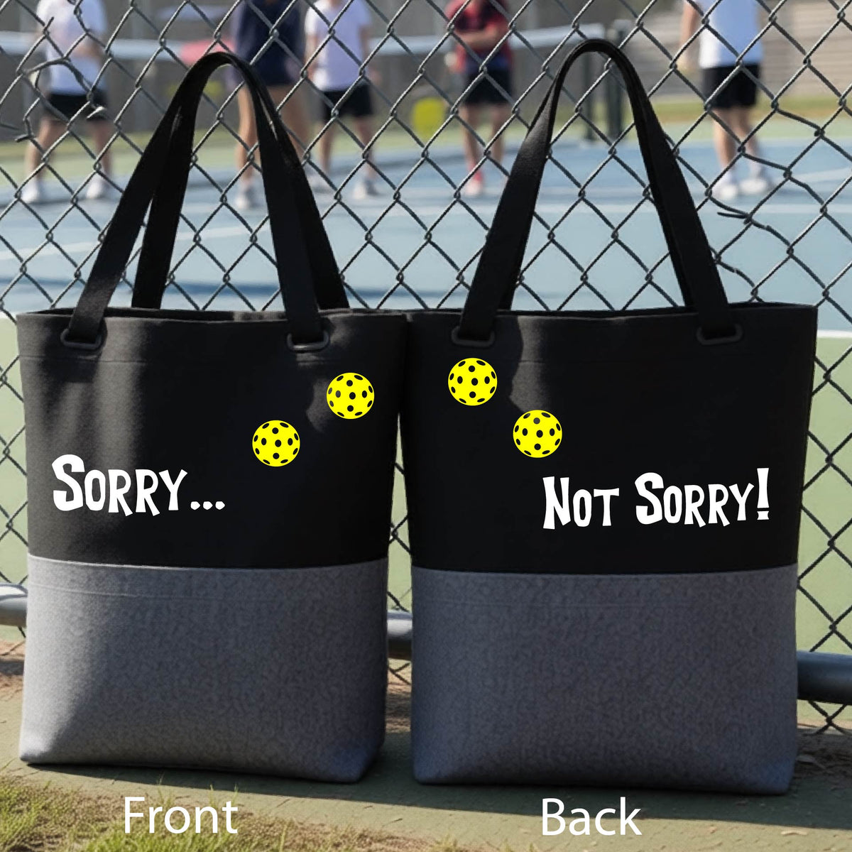 A close-up of a gray and black medium size duffel bag hanging on a fence at a pickleball court, with pickleball motif design on the front and brim and features the playful word “SORRY…” in bold white lettering, accented a colorful pickleball, with the words 'Not sorry" curved underneath on the front of the duffel bag. 