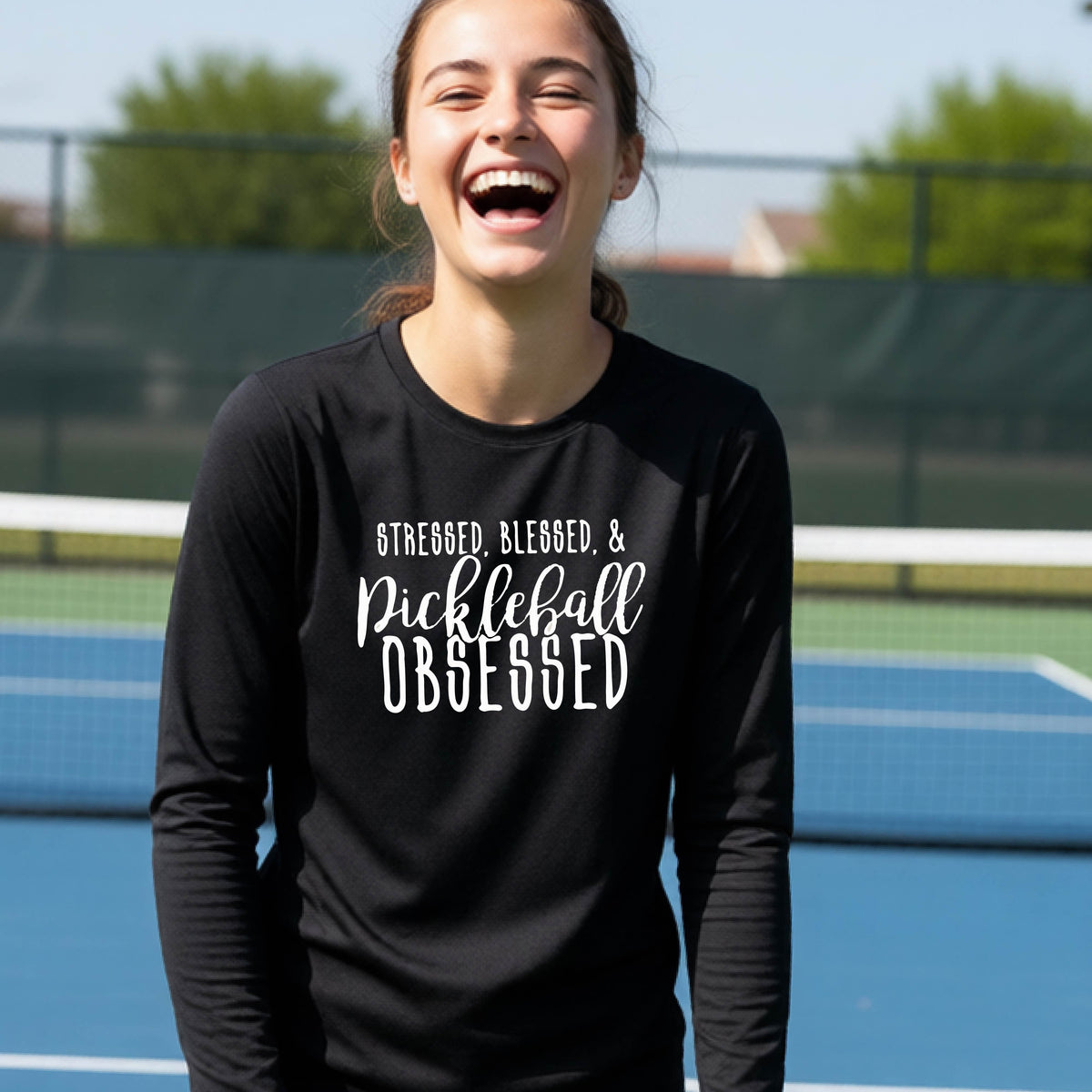 A young person on a pickleball court wearing a black long-sleeve t-shirt with a white graphic of the words “Stressed, Blessed & Pickleball Obsessed” on the front of the shirt.