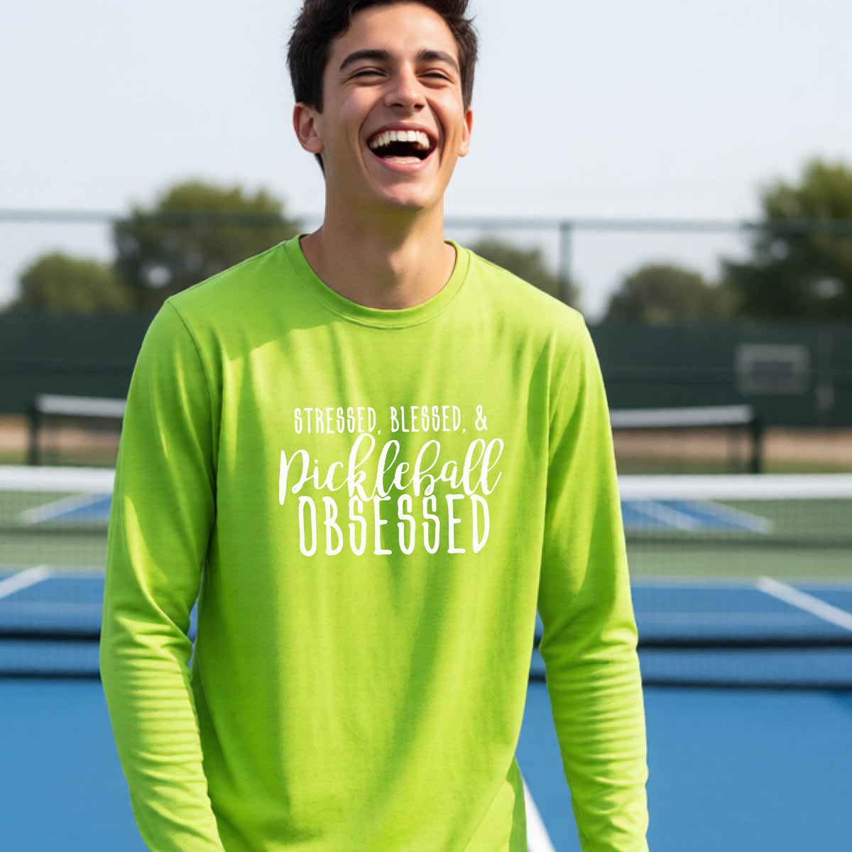 A young person on a pickleball court wearing a lime green long-sleeve t-shirt with a white graphic of the words “Stressed, Blessed & Pickleball Obsessed” on the front of the shirt.