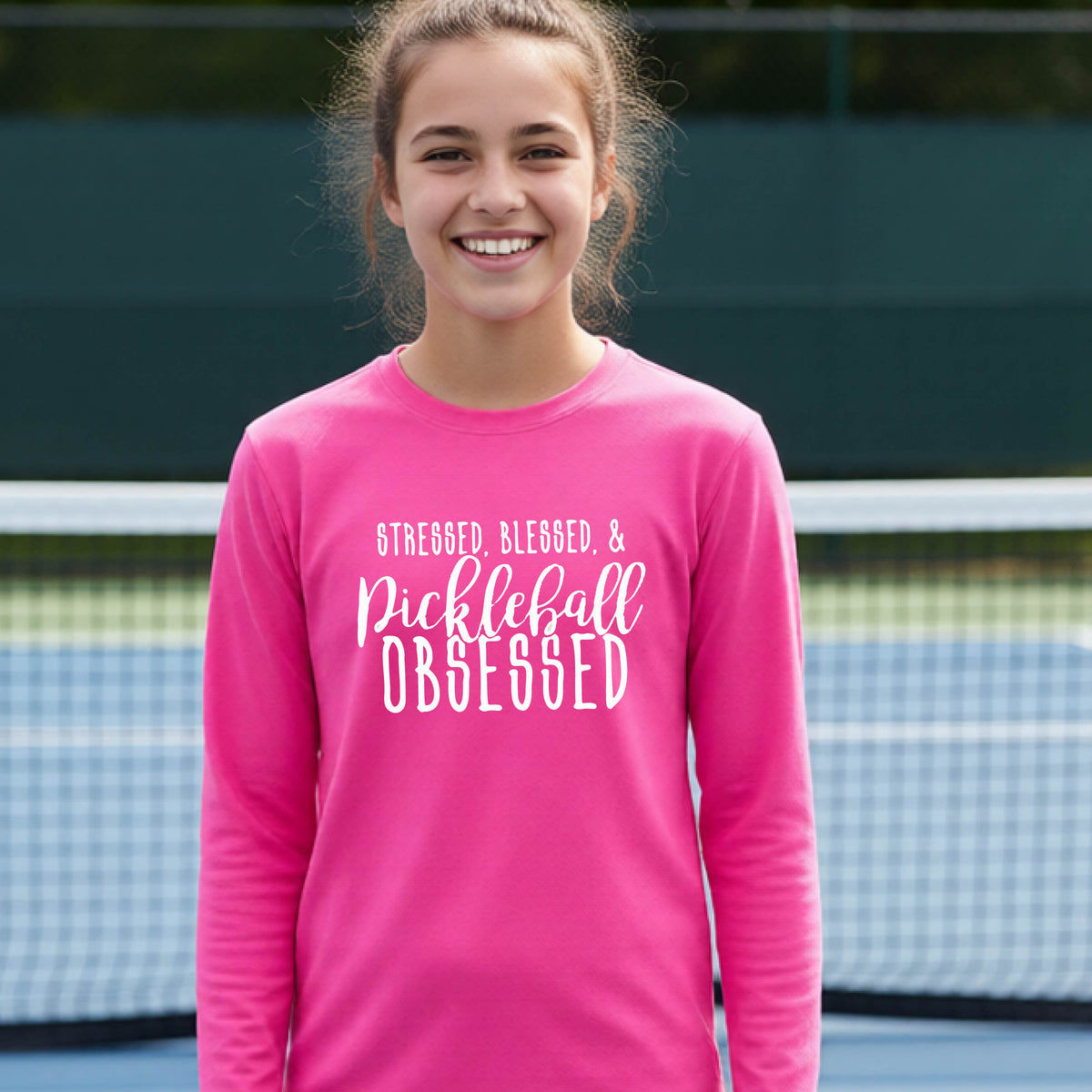 A young person on a pickleball court wearing a pink long-sleeve t-shirt with a white graphic of the words “Stressed, Blessed & Pickleball Obsessed” on the front of the shirt.