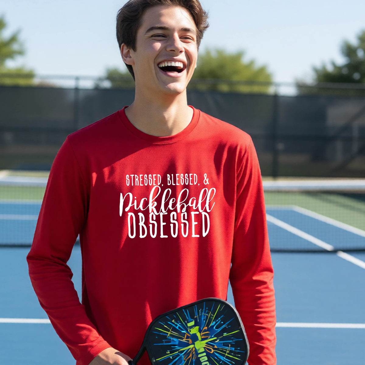 A young person on a pickleball court wearing a red long-sleeve t-shirt with a white graphic of the words “Stressed, Blessed & Pickleball Obsessed” on the front of the shirt.