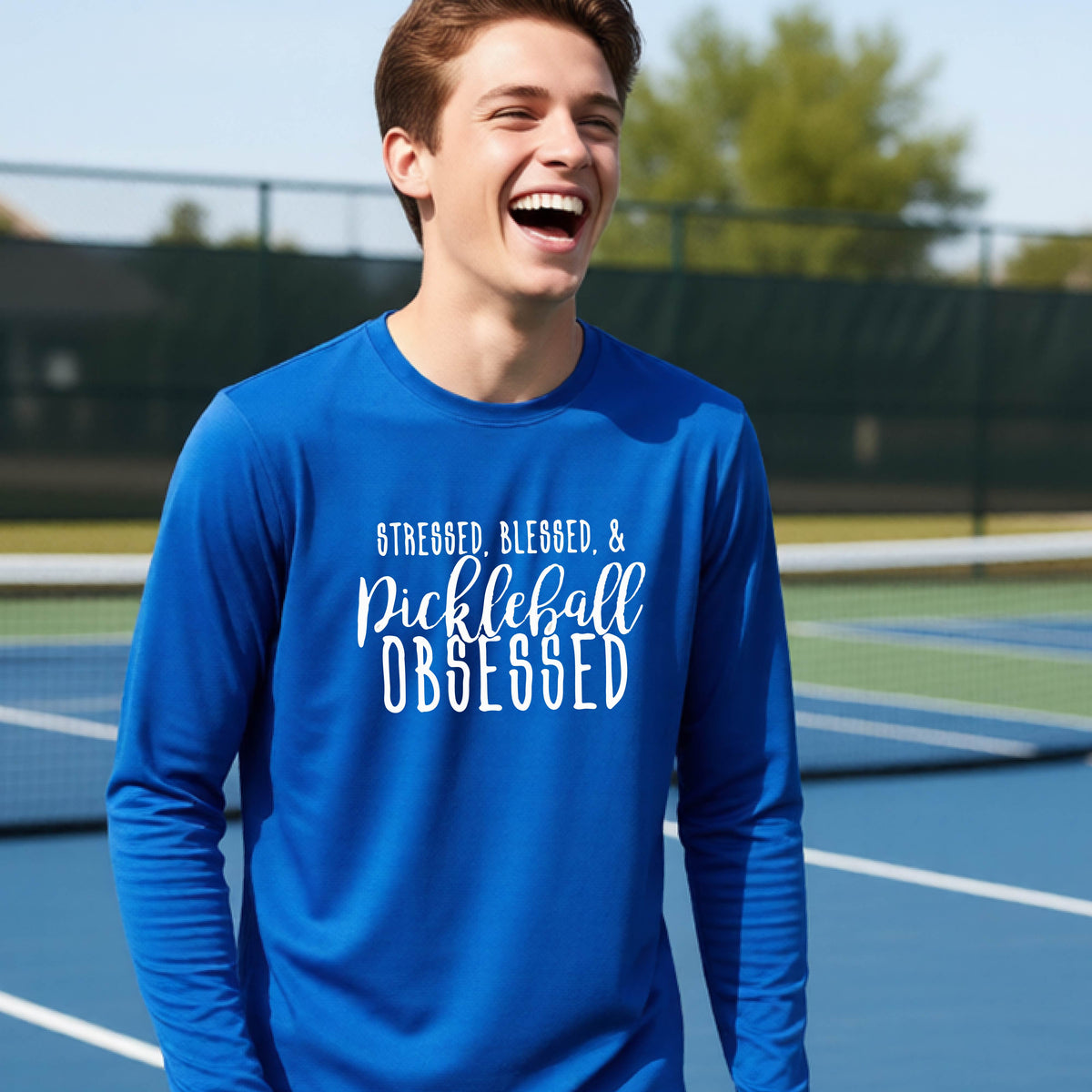 A young person on a pickleball court wearing a royal blue long-sleeve t-shirt with a white graphic of the words “Stressed, Blessed & Pickleball Obsessed” on the front of the shirt.