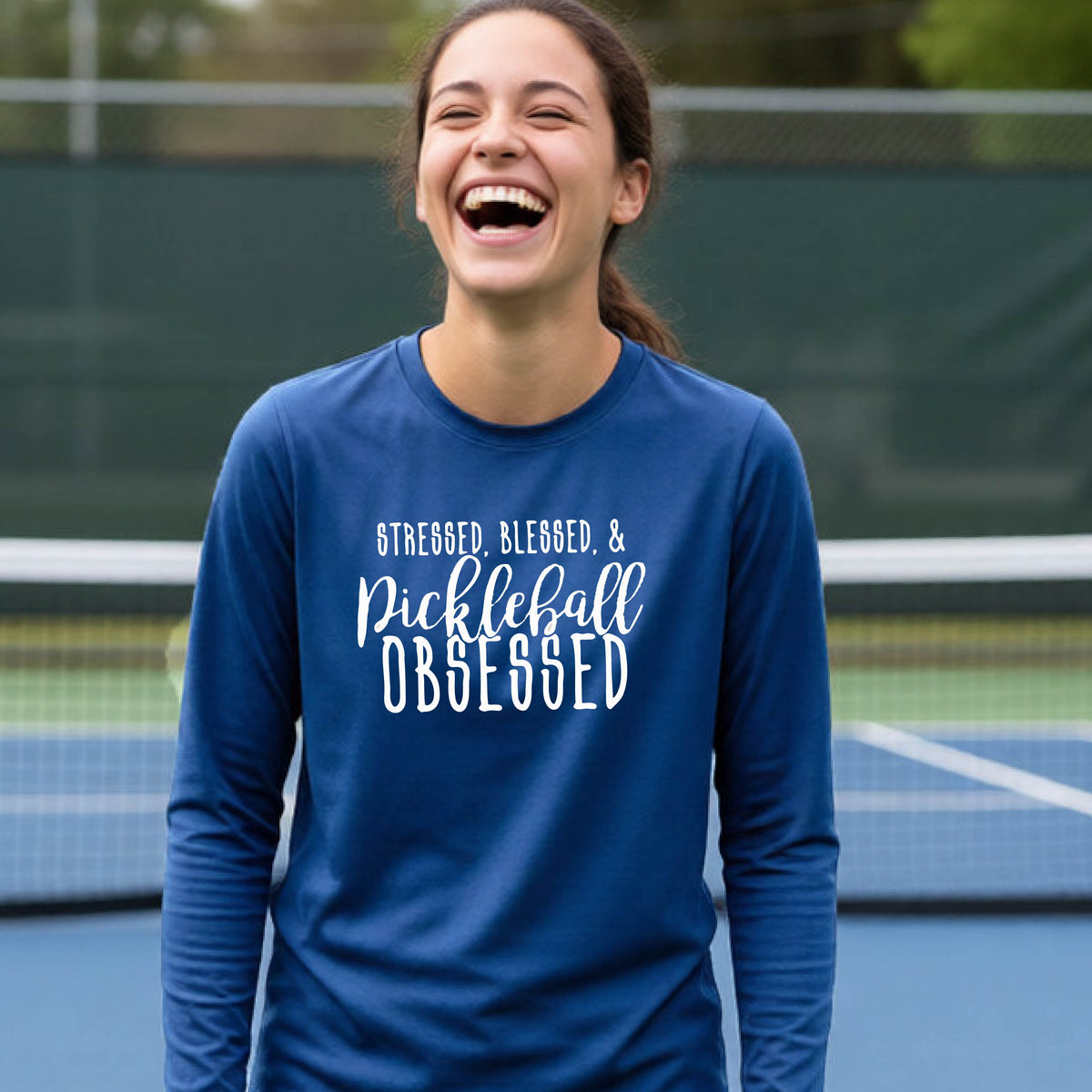 A young person on a pickleball court wearing a royal blue long-sleeve t-shirt with a white graphic of the words “Stressed, Blessed & Pickleball Obsessed” on the front of the shirt.
