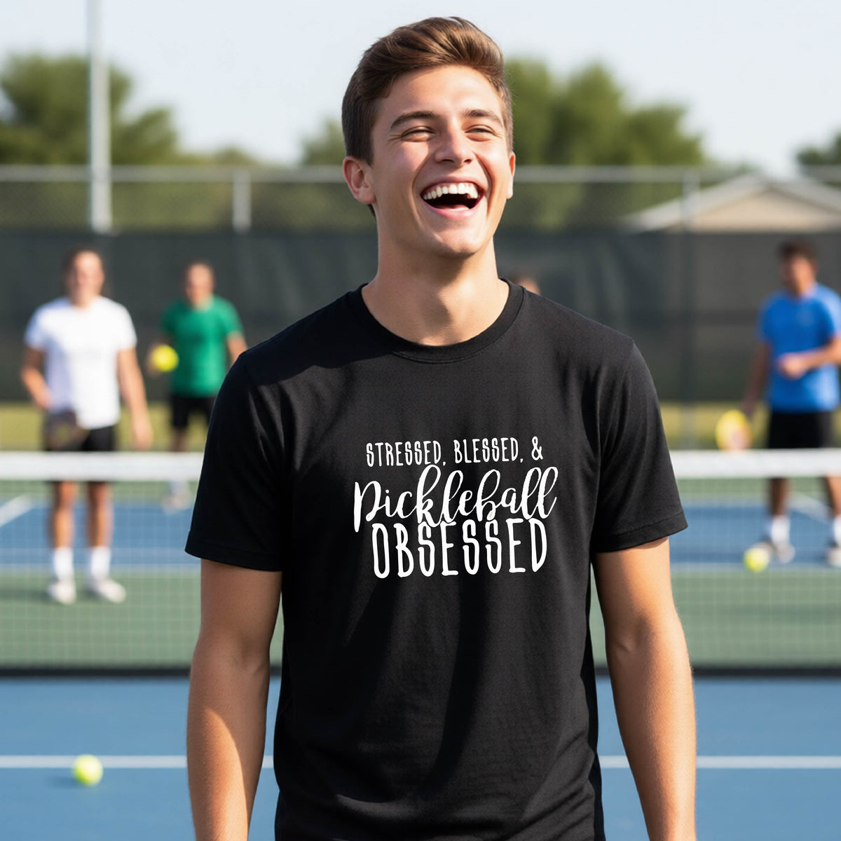 A young person on a pickleball court wearing a black short-sleeve t-shirt with a white graphic of the words “Stressed, Blessed & Pickleball Obsessed” on the front of the shirt.