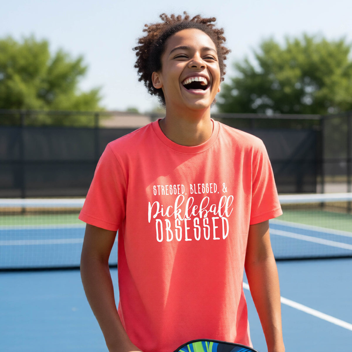 A young person on a pickleball court wearing a coral pink short-sleeve t-shirt with a white graphic of the words “Stressed, Blessed & Pickleball Obsessed” on the front of the shirt.