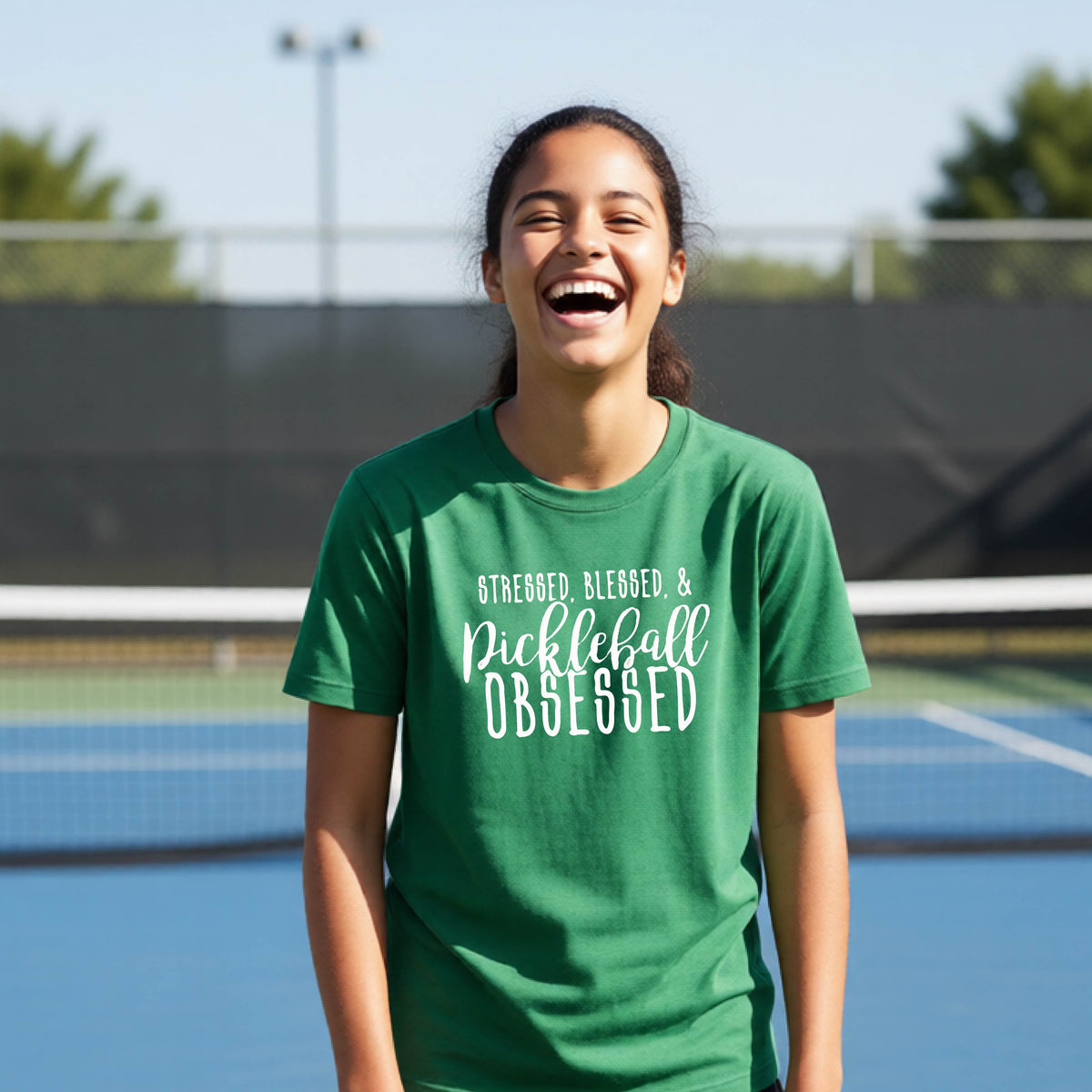 A young person on a pickleball court wearing a green short-sleeve t-shirt with a white graphic of the words “Stressed, Blessed & Pickleball Obsessed” on the front of the shirt.