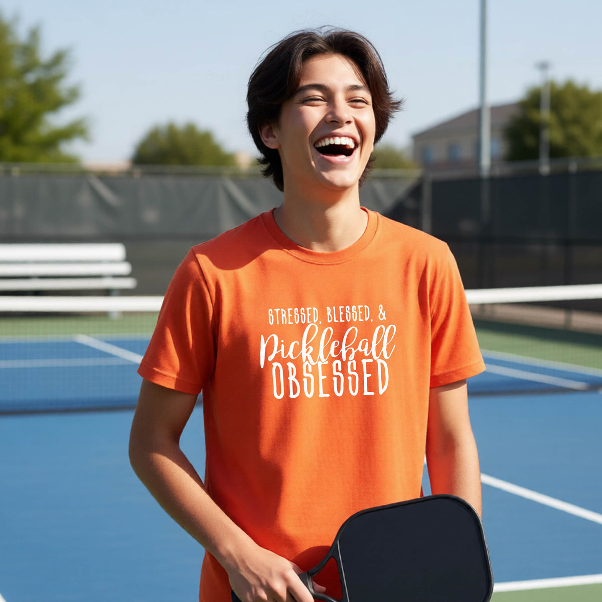 A young person on a pickleball court wearing an orange short-sleeve t-shirt with a white graphic of the words “Stressed, Blessed & Pickleball Obsessed” on the front of the shirt.