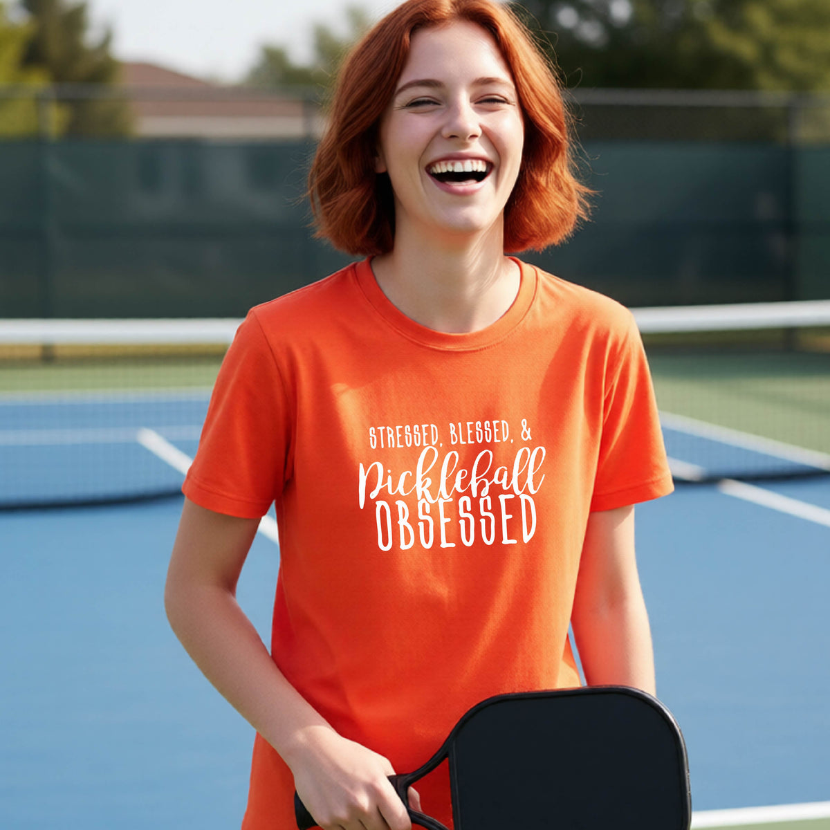 A young person on a pickleball court wearing an orange short-sleeve t-shirt with a white graphic of the words “Stressed, Blessed & Pickleball Obsessed” on the front of the shirt.