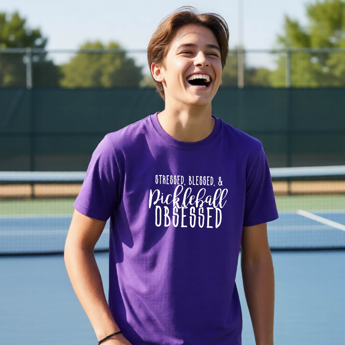 A young person on a pickleball court wearing a purple short-sleeve t-shirt with a white graphic of the words “Stressed, Blessed & Pickleball Obsessed” on the front of the shirt.