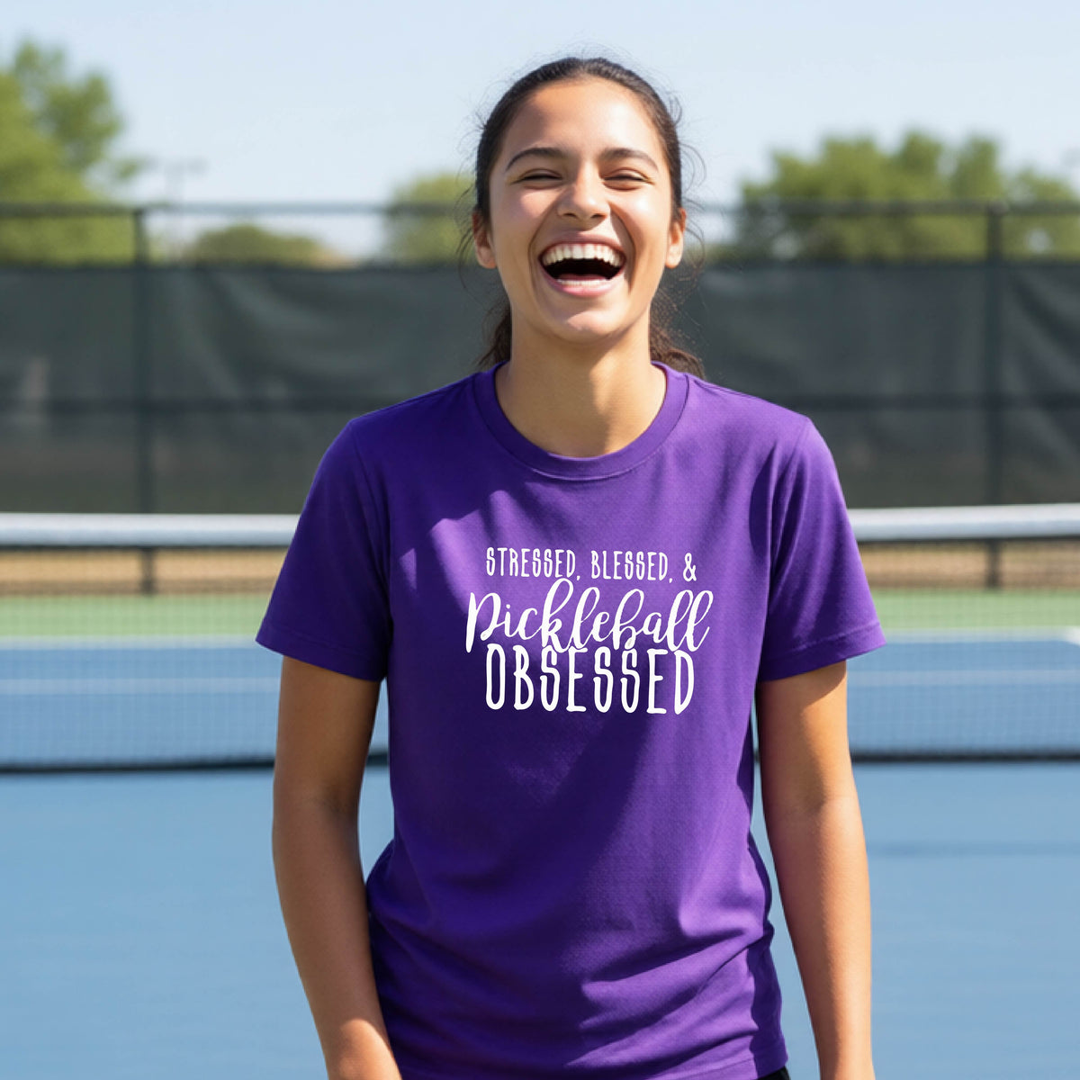 A young person on a pickleball court wearing a purple short-sleeve t-shirt with a white graphic of the words “Stressed, Blessed & Pickleball Obsessed” on the front of the shirt.