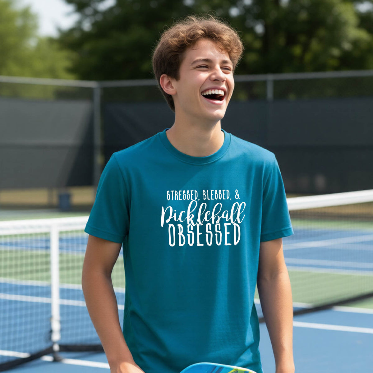 A young person on a pickleball court wearing a teal short-sleeve t-shirt with a white graphic of the words “Stressed, Blessed & Pickleball Obsessed” on the front of the shirt.