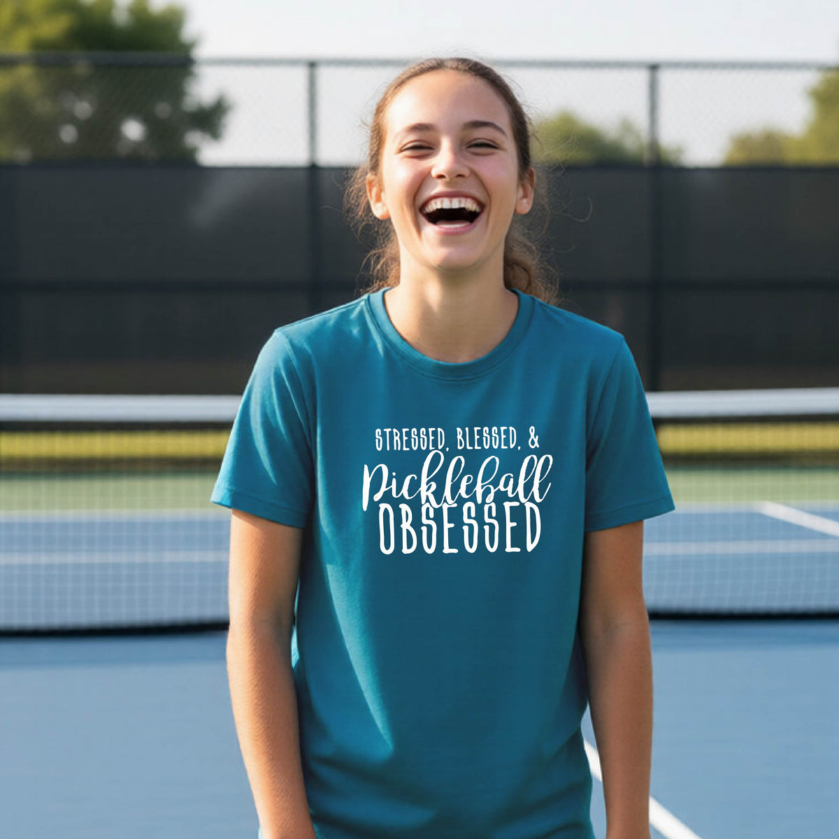 A young person on a pickleball court wearing a teal short-sleeve t-shirt with a white graphic of the words “Stressed, Blessed & Pickleball Obsessed” on the front of the shirt.