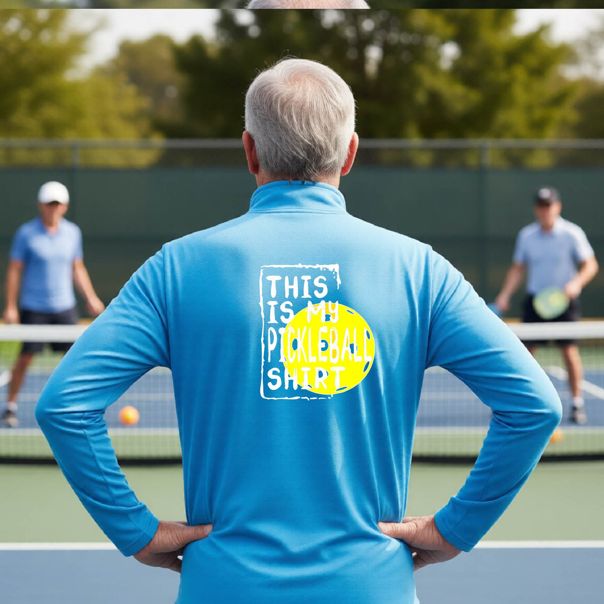 A man on a pickleball court seen from behind wearing a bright blue long-sleeve shirt with a graphic on the back that reads 'THIS IS MY PICKLEBALL SHIRT' in white text over a yellow pickleball.