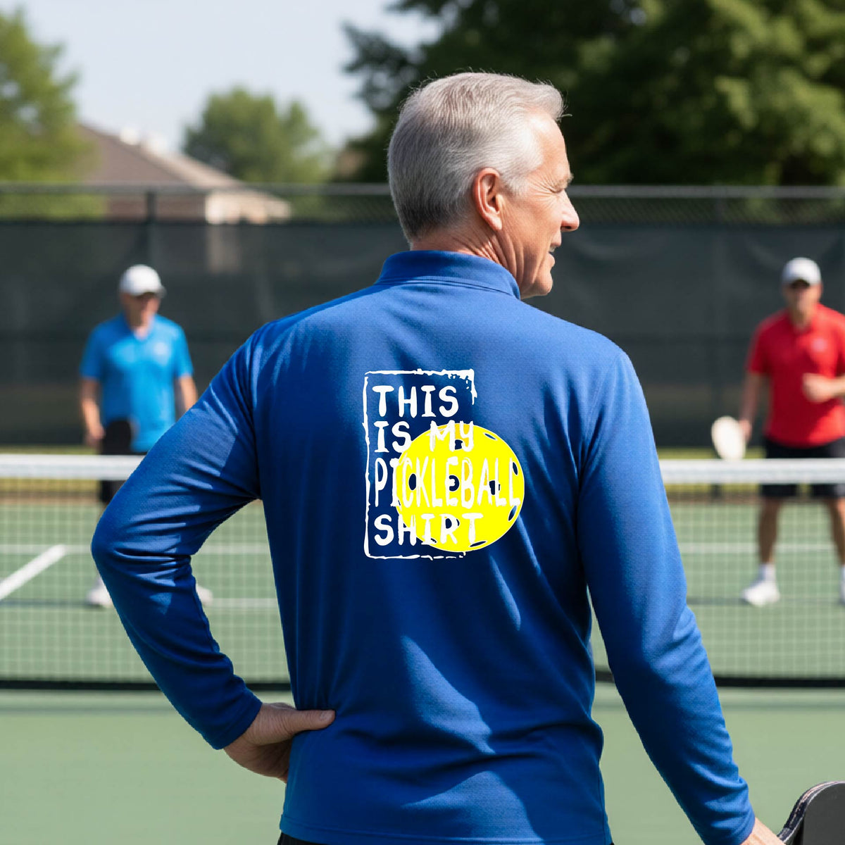 A man on a pickleball court seen from behind wearing a royal blue long-sleeve shirt with a graphic on the back that reads 'THIS IS MY PICKLEBALL SHIRT' in white text over a yellow pickleball.