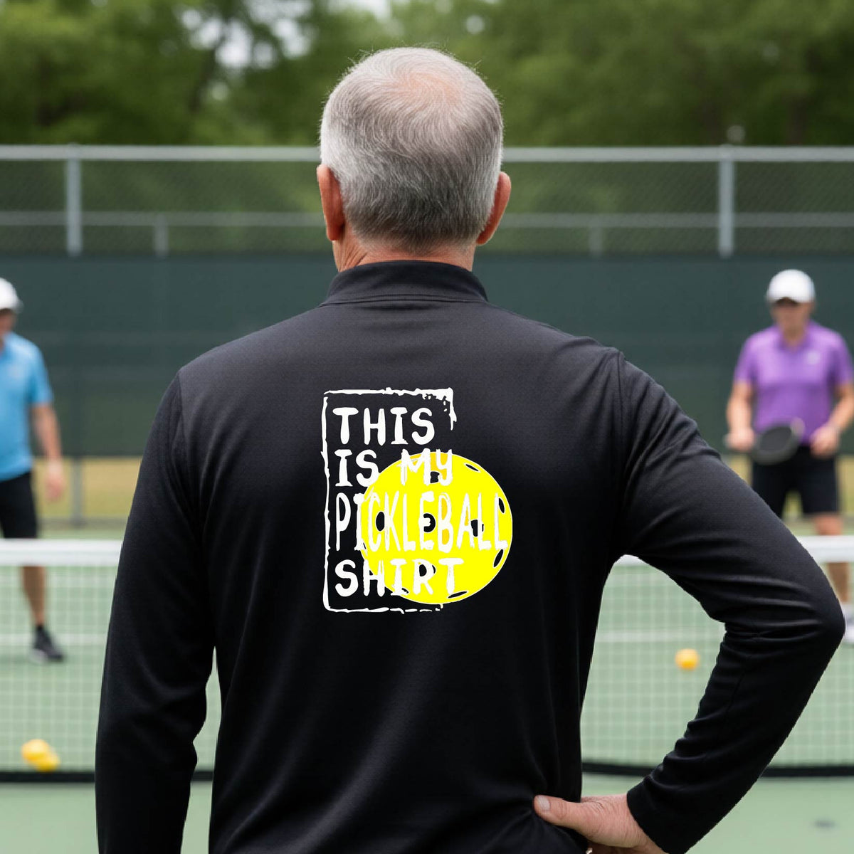 A man on a pickleball court seen from behind wearing a black  long-sleeve shirt with a graphic on the back that reads 'THIS IS MY PICKLEBALL SHIRT' in white text over a yellow pickleball.