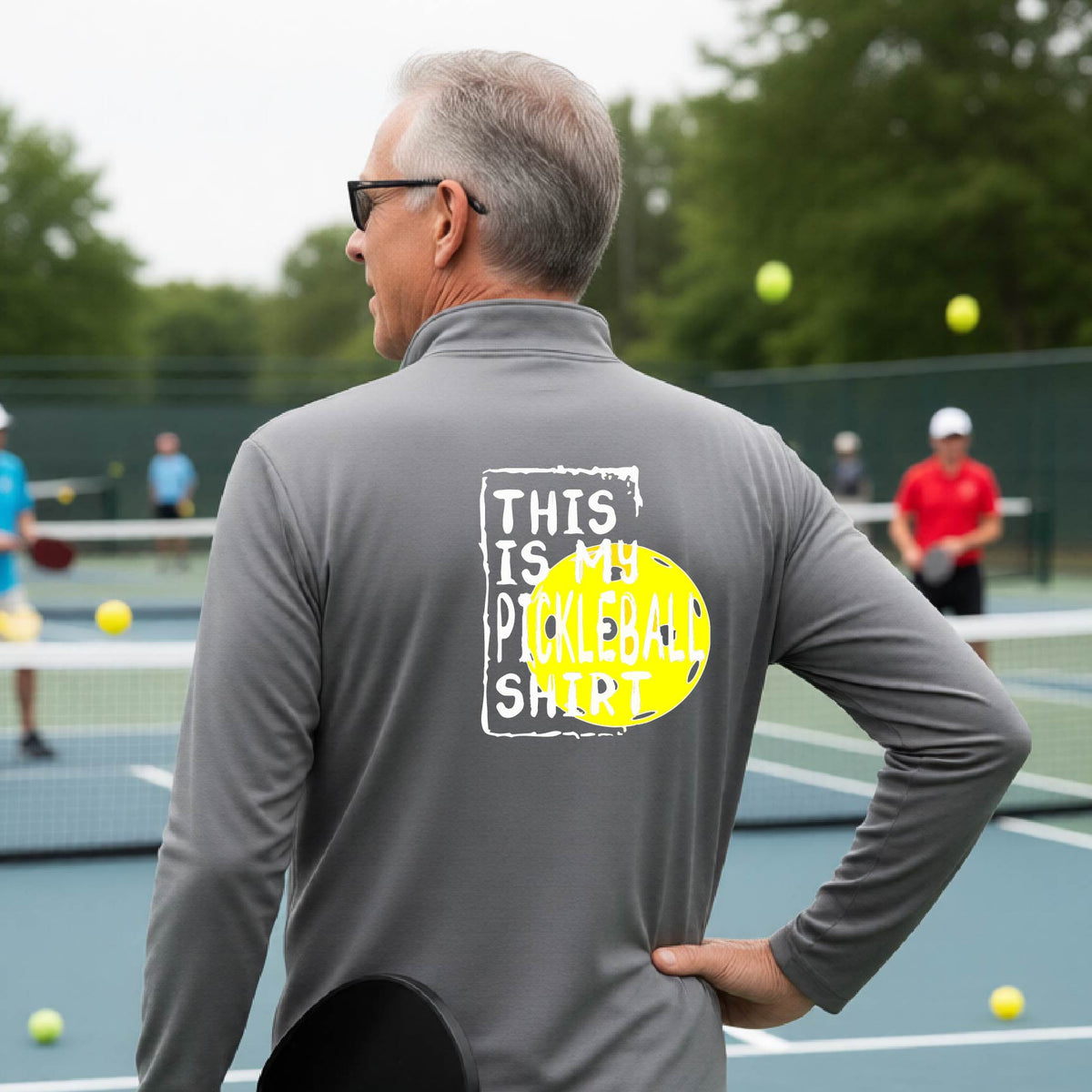 A man on a pickleball court seen from behind wearing a gray long-sleeve shirt with a graphic on the back that reads 'THIS IS MY PICKLEBALL SHIRT' in white text over a yellow pickleball.