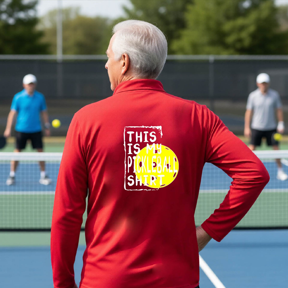 A man on a pickleball court seen from behind wearing a bright red long-sleeve shirt with a graphic on the back that reads 'THIS IS MY PICKLEBALL SHIRT' in white text over a yellow pickleball.