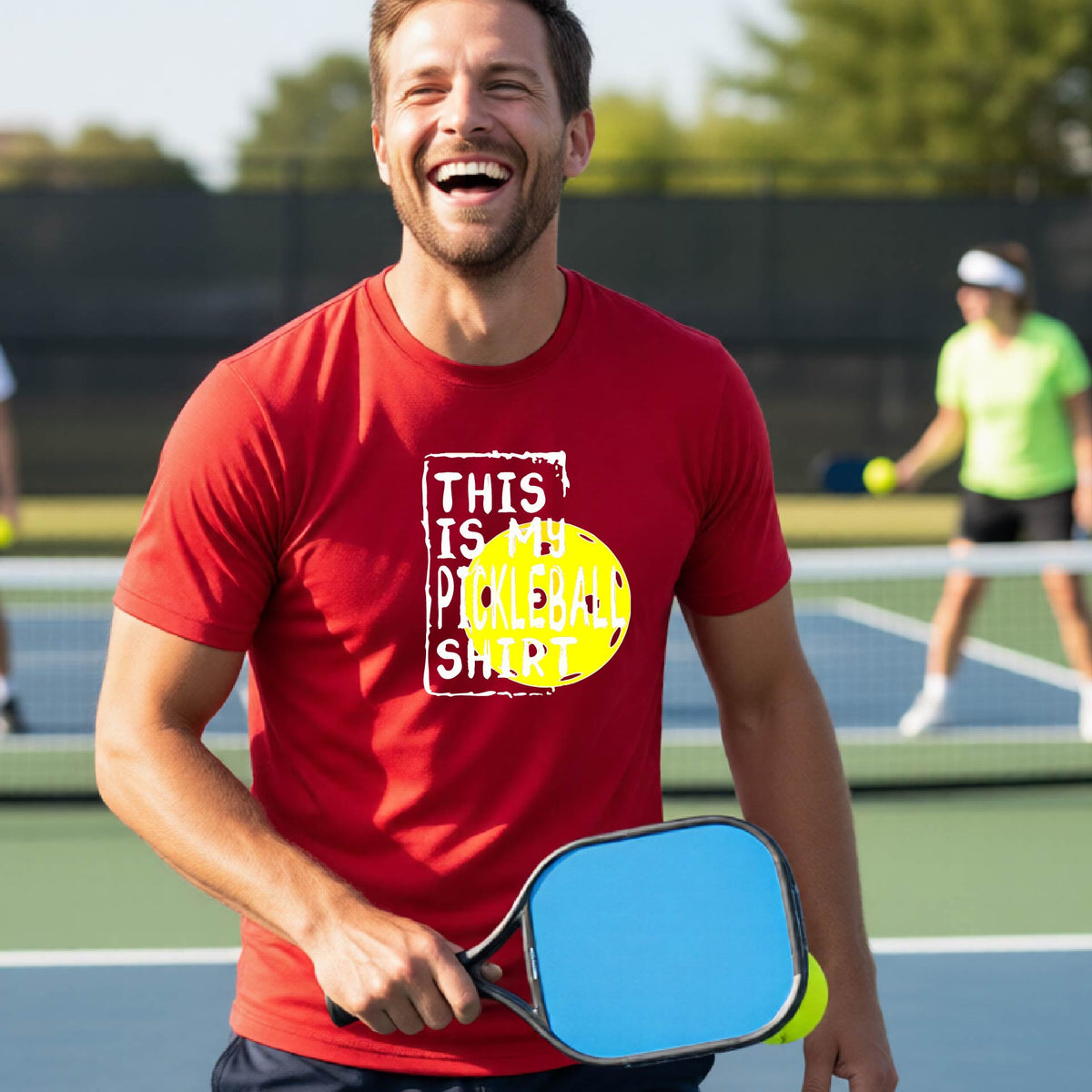 A man on an outdoor pickleball court wearing a red t-shirt with a graphic that reads 'THIS IS MY PICKLEBALL SHIRT' in white with a yellow pickleball icon. 
