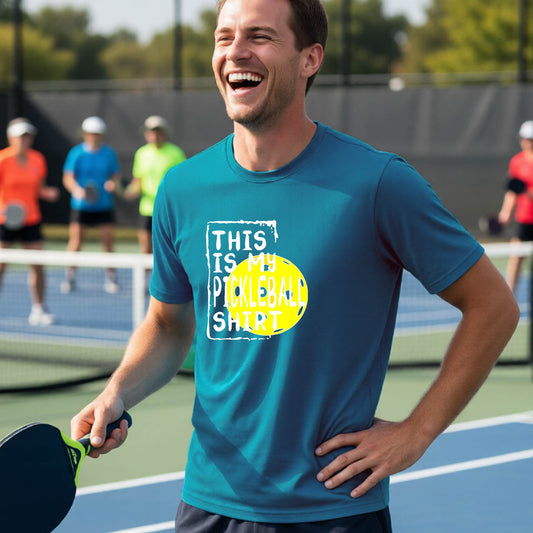 A man on an outdoor pickleball court wearing a teal t-shirt with a graphic that reads 'THIS IS MY PICKLEBALL SHIRT' in white with a yellow pickleball icon. 