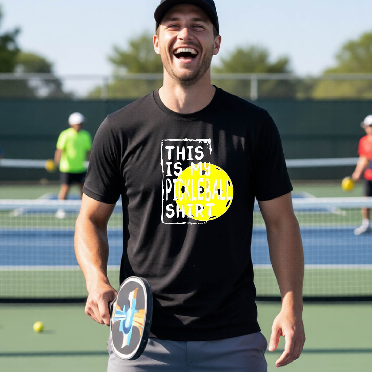 A man on an outdoor pickleball court wearing a black t-shirt with a graphic that reads 'THIS IS MY PICKLEBALL SHIRT' in white with a yellow pickleball icon. 