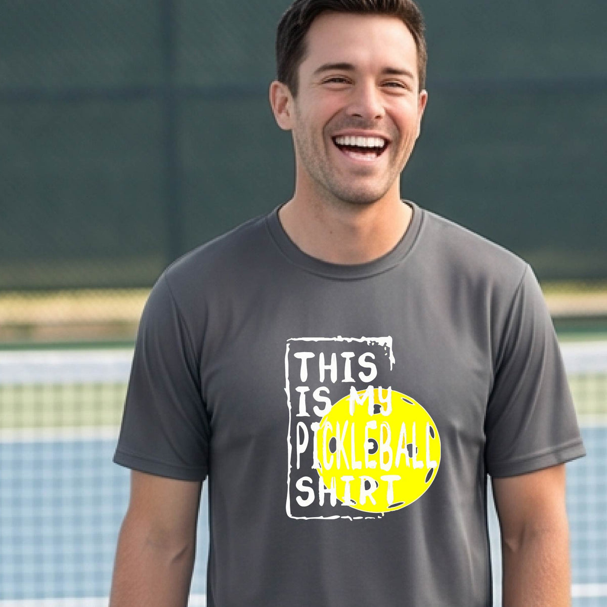 A man on an outdoor pickleball court wearing a gray t-shirt with a graphic that reads 'THIS IS MY PICKLEBALL SHIRT' in white with a yellow pickleball icon. 