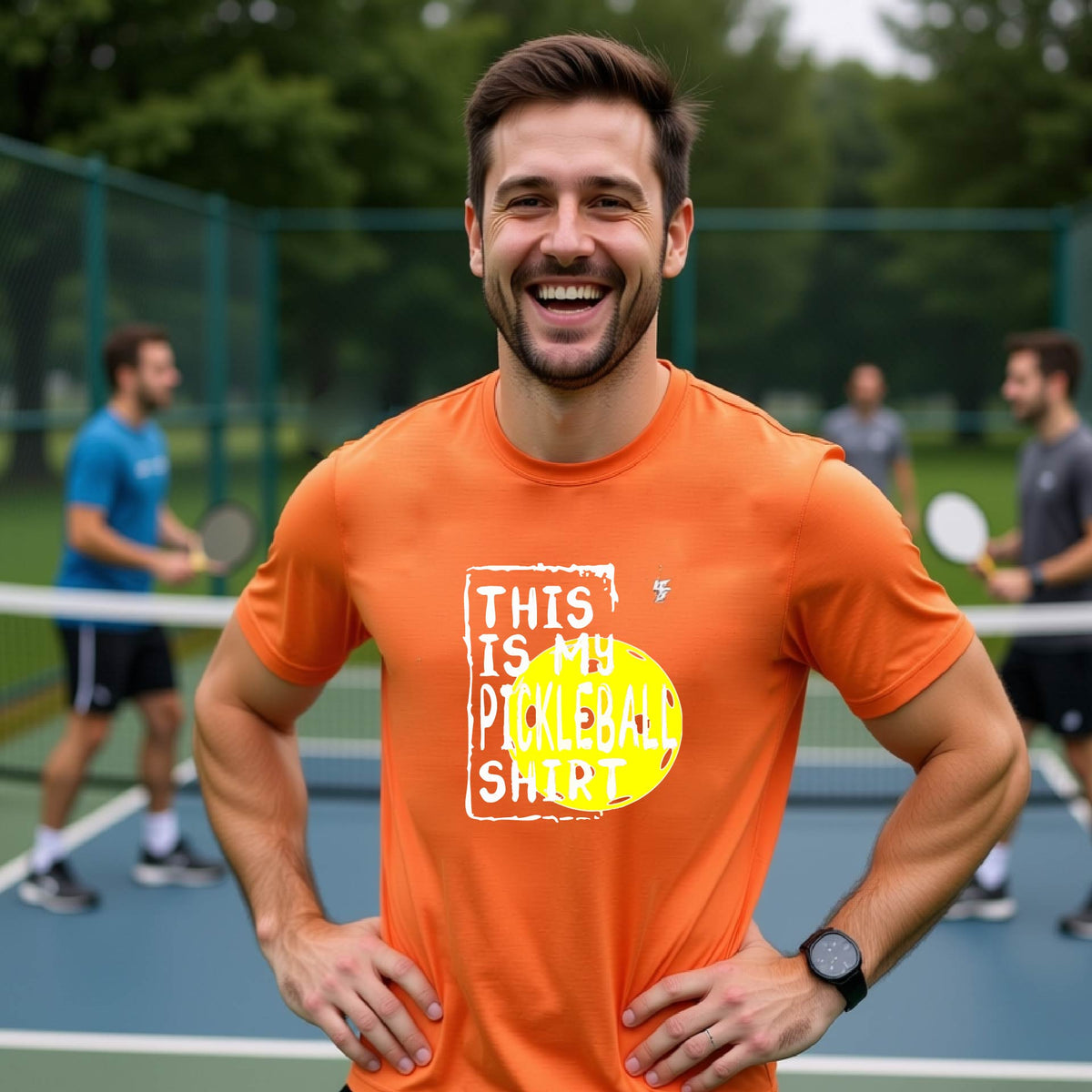 A man on an outdoor pickleball court wearing an orange t-shirt with a graphic that reads 'THIS IS MY PICKLEBALL SHIRT' in white with a yellow pickleball icon. 