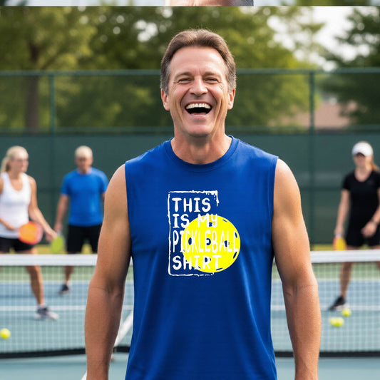 A man on a pickleball court wearing a blue sleeveless shirt with a yellow and white graphic that reads 'THIS IS MY PICKLEBALL SHIRT' and a yellow pickleball icon.