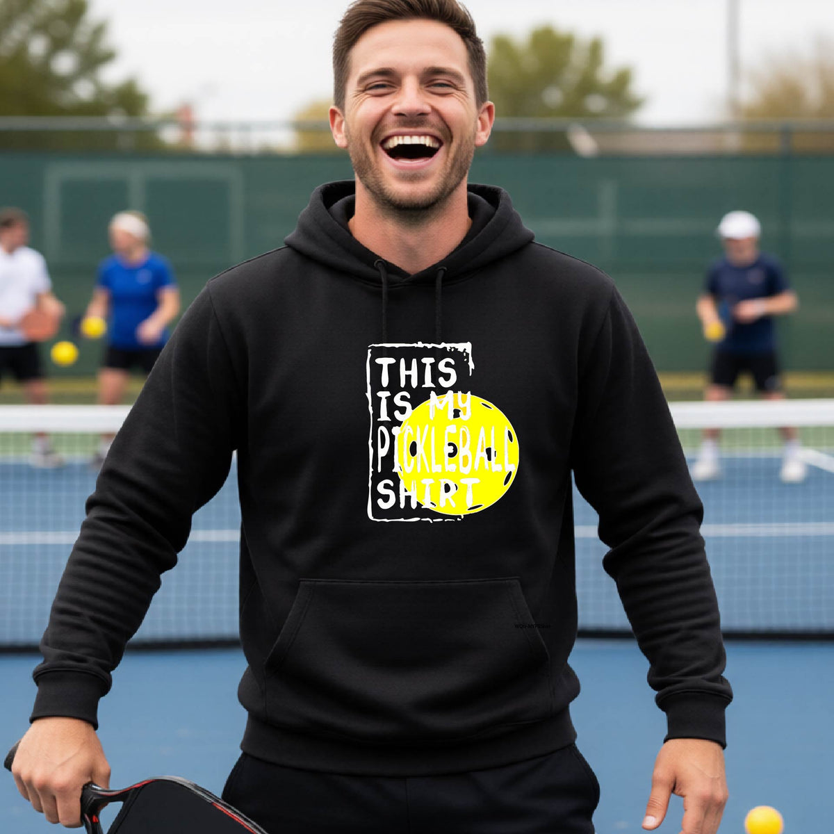A man wearing a black hoodie with a yellow and white graphic that reads 'THIS IS MY PICKLEBALL SHIRT' stands on a pickleball court. 