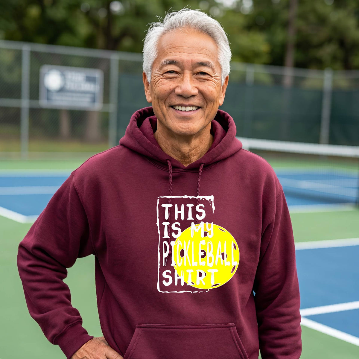A man wearing a maroon hoodie with a yellow and white graphic that reads 'THIS IS MY PICKLEBALL SHIRT' stands on a pickleball court. 