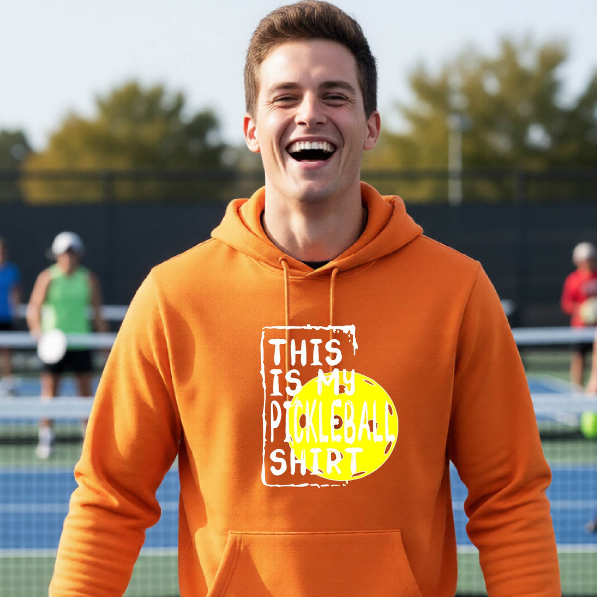 A man wearing an orange hoodie with a yellow and white graphic that reads 'THIS IS MY PICKLEBALL SHIRT' stands on a pickleball court. 