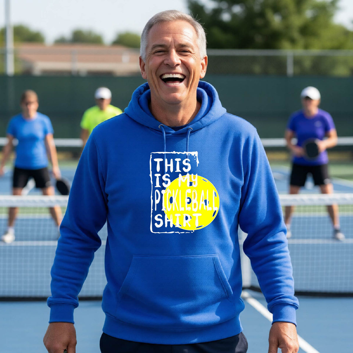 A man wearing a royal blue hoodie with a yellow and white graphic that reads 'THIS IS MY PICKLEBALL SHIRT' stands on a pickleball court. 